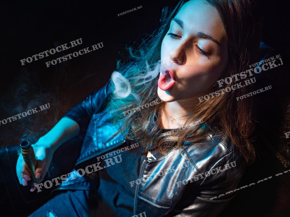 Фотография изображает молодую женщину в кожаной куртке, которая сидит и выпускает кольца дыма от электронной сигареты. Снимок выполнен в неоновом свете, он создает атмосферу ночной жизни и современного стиля.